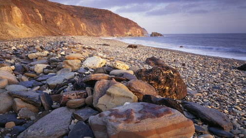 A stony beach; low morning sunlight casts an orange glow onto the face of cliffs to the left and larger rocks in the foreground. The sea to the right is dark blue-grey and thin clouds above have a purple tinge.